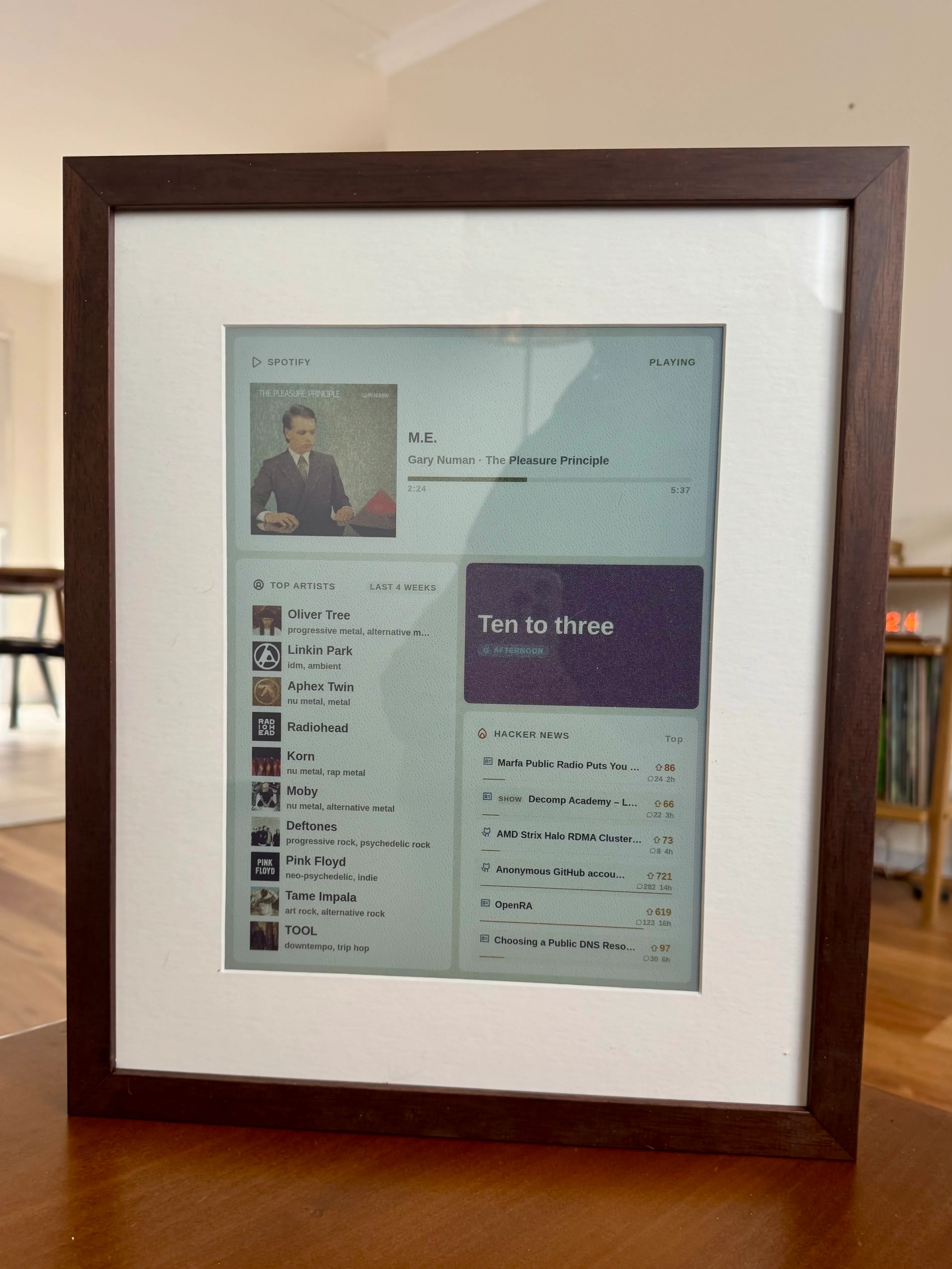 A framed portrait e-paper panel showing a now-playing dashboard and Hacker News