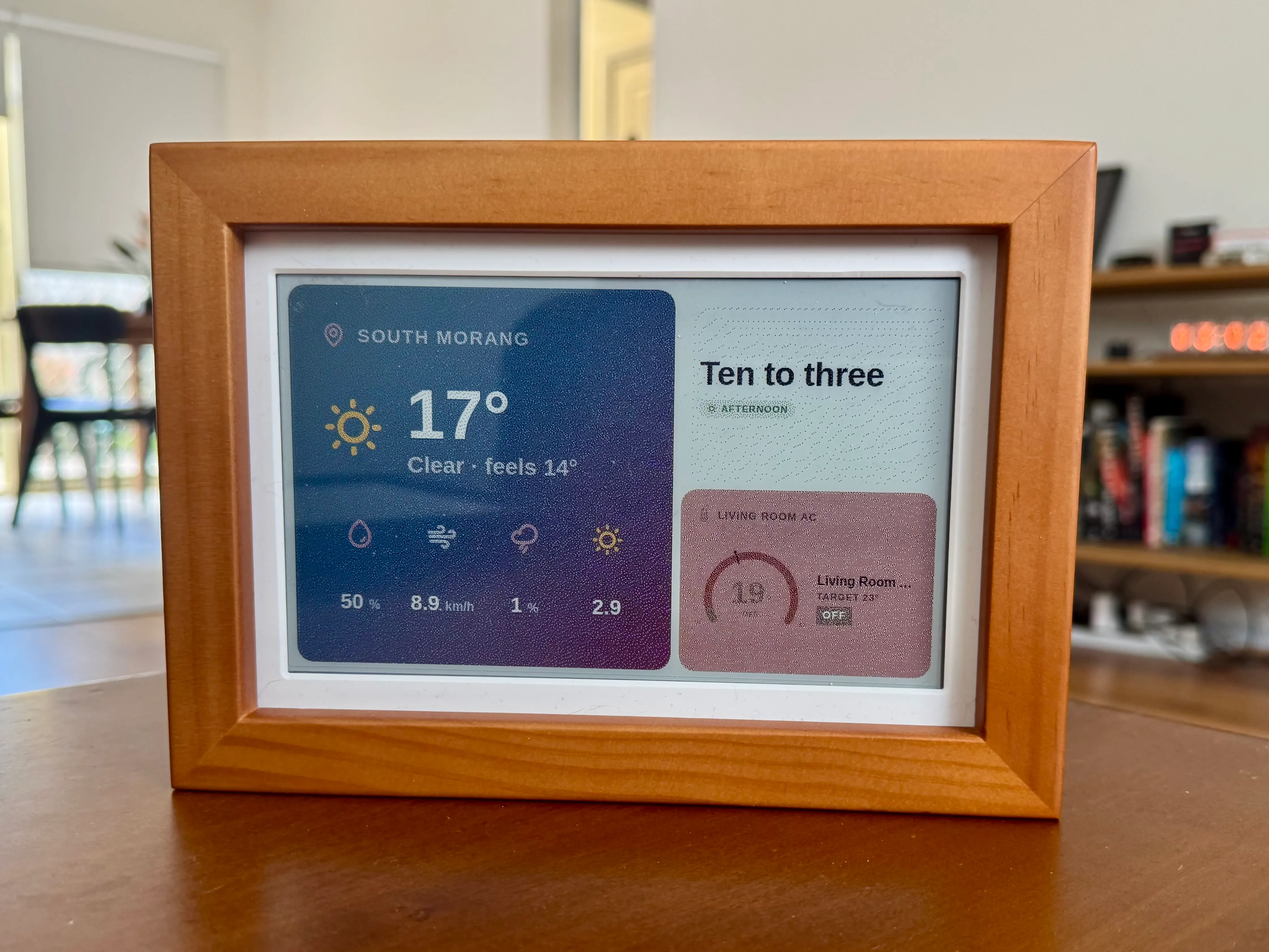 A wood-framed e-paper panel showing weather, a word clock and climate on a desk